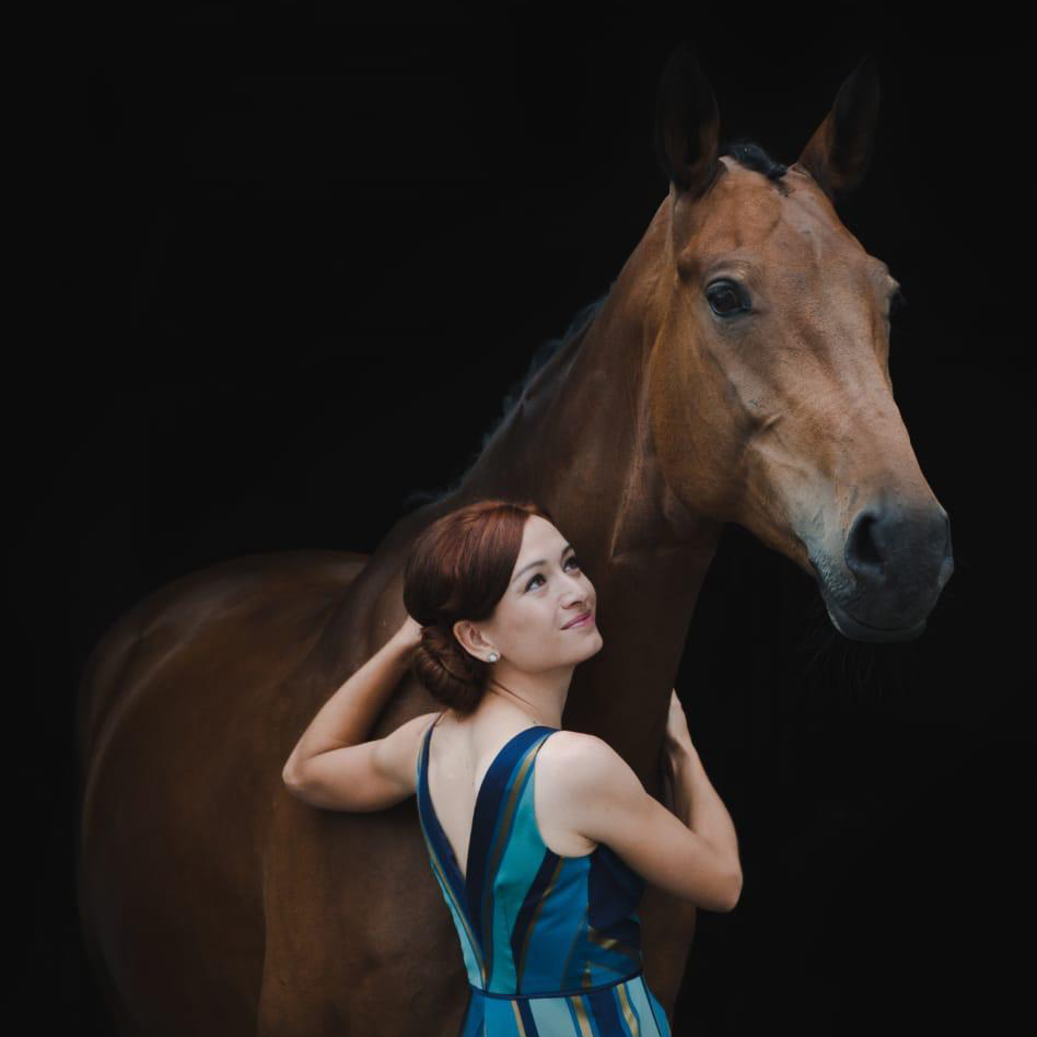 A woman in a blue and teal striped sleeveless dress with her dark hair in a bun poses next to a chestnut brown horse against a black background, looking up at the horse while touching her hair with one hand in an elegant portrait-style photograph.